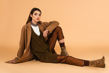 young woman in brown coat, satin pants and leather shoes sitting on beige background