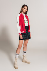 young woman in stylish jacket, plaid skirt and high boots posing on grey background
