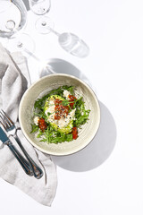 Avocado and stracciatella cheese salad. Fancy dinning with Italian salad bowl on white table with simple contemporary decor. Sunlight and harsh shadow still life. Avocado, cheese and tomato salad.