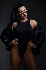 beautiful woman with a sports figure in a black bodysuit, black jacket and sunglasses posing in the studio on a gray background 
