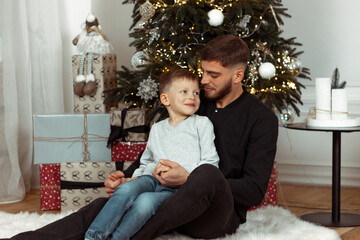 Father with son smiling, hugging, and sitting near the Christmas tree in a cozy decorated room for winter holidays. Family festive portraits. New Year celebrations. Winter magic time, fairytale vibes.