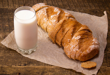 A sliced rye loaf lying on a wooden table and a glass of milk.Bread lying on paper next to an old knife and milk.