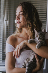 Portrait of  relaxed young female model holding cat  , with natural makeup, wavy hair, dressed in white clothes on background .People, beauty, face care - 450351709