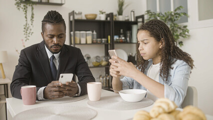 Black gadget-addicted father and daughter holding smartphones during breakfast