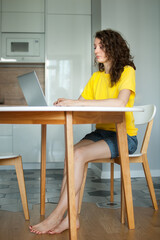 Young woman with curly hair and yellow shirt is working from home using her laptop at the kitchen table in her apartment, remote work, freelance