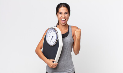 young hispanic woman feeling shocked,laughing and celebrating success and holding a weight scale....