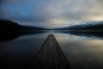 立ち込む雲が映りこんだ静かな朝の湖に伸びる一本の木製桟橋