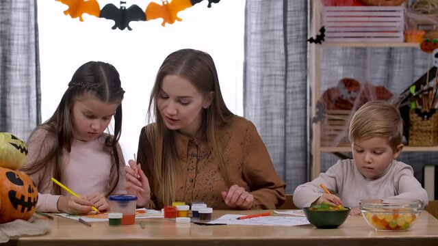 Happy caucasian woman and her two children, daughter and son, are sitting at the table, talking and drawing pumpkins together. While mom is distracted, son is eating treats. Slow motion. Close up.
