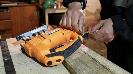 repairing the cord of an electric jigsaw at the workplace by the hands of a master, repairing a tool on a tree by a carpenter in uniform, men's hands repairing the wire of an orange jigsaw