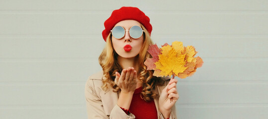Autumn portrait of beautiful young woman model with yellow maple leaves blowing her lips with lipstick wearing a red french beret on gray background