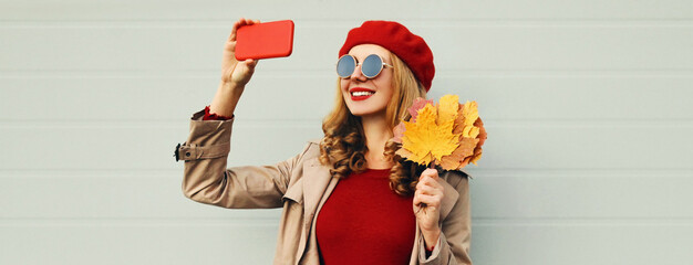 Autumn portrait of beautiful smiling woman taking selfie by smartphone with yellow maple leaves wearing a red french beret on gray background