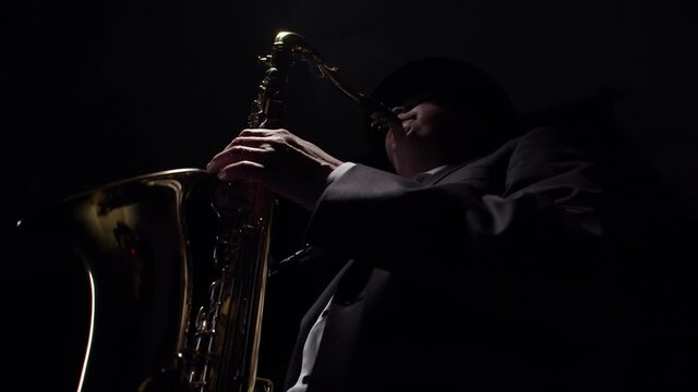 Male saxophonist playing a saxophone with hands and mouth is caught in the light from above in a darkness studio. Concept of classical music.