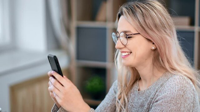 Happy young freelancer female chatting surfing internet use smartphone at workplace enjoying break