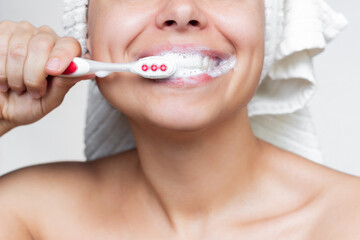 Cropped shot of a young smiling woman with a white towel on her head after a shower is brushing her teeth with a toothbrush on a white background. Morning and evening routine. Close up