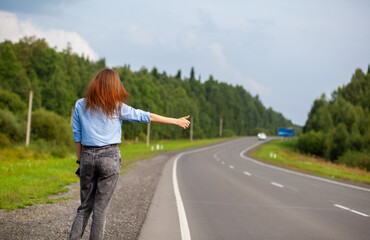 Obraz premium The girl stops the car on the highway with her hand. Stylish woman on the road stops the car go on a journey. A road in the middle of the forest.