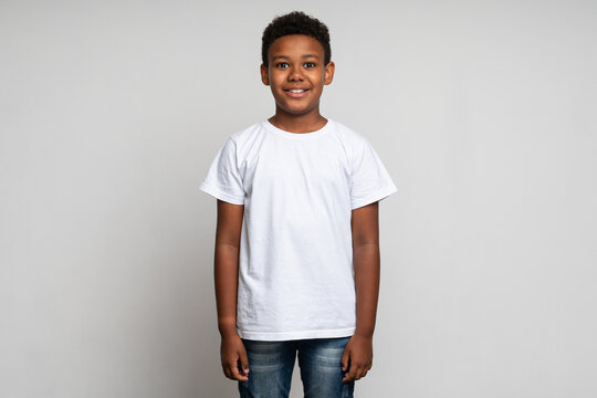 Horizontal view of cute little boy with stylish curly hairdo in white T-shirt standing and looking at camera with happy face. Indoor studio shot isolated on white background