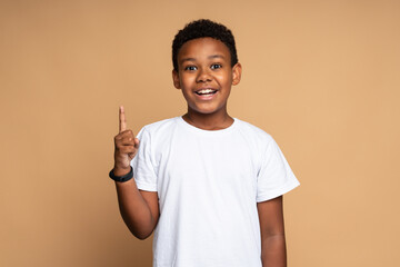 Portrait of amazed cute little boy with curly hair pointing to empty place on background. Surprised preschooler showing copy space for promotional ad. Indoor studio shot