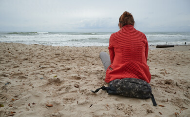 Turystka siedzi na plecaku na brzegu morza i patrzy w dal na delikatne fale. Szum jesiennego morza. - obrazy, fototapety, plakaty