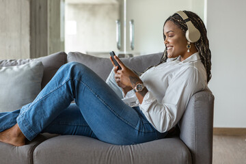 Happy young woman listening to music with headphones and using mobile phone while leaning on a sofa at home
