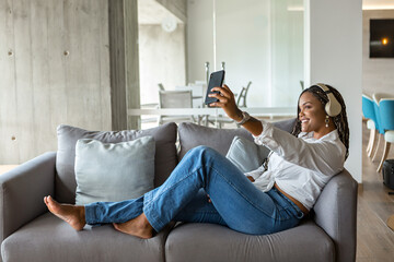 Happy young woman listening to music with headphones and using mobile phone to take a selfie while leaning on a sofa at home