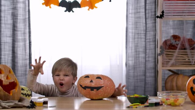 In a room decorated for Halloween, the boy hid under a table with a pumpkin with a scary face painted on it. The child jumps out from under the table and begins to scare. Slow motion. Close up.