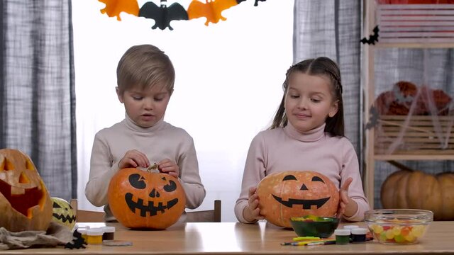 Two children, a boy and a girl, raise pumpkins in front of them with scary faces painted. They put pumpkins on the table and make faces. Kids love Halloween. Slow motion. Close up.