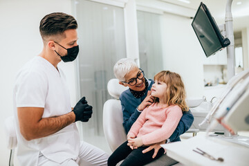 Cute little girl sitting on dental chair and having dental treatment. Her grandmother is with her. Dentist is wearing protective face mask due to coronavirus pandemic.