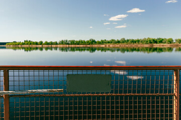 A fence on board the ferry with a sign with a view of a summer landscape with a river, a blue sky with clouds, and the shore