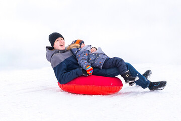 Fototapeta na wymiar A cheerful young man and a little boy ride on a snow slide on a cheesecake on a winter day