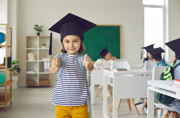 Portrait of elementary schoolgirl in square academic cap standing in classroom. Indoor shot of happy school student in mortarboard. Cheerful smiling little child looking at camera and giving thumbs up