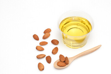 Yellow almond oil in a clear bowl with a wooden spoon next to it and several almonds on a white background