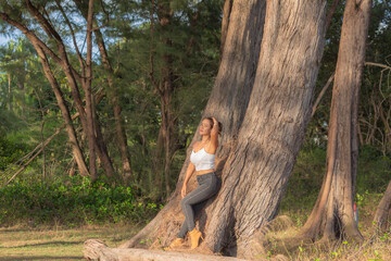 woman wearing sexy white tank top Wearing black tights and boots, .standing leaning against a large pine tree. .Tree Tunnel beside the beach. Large pine trees line up to form a tunnel background.