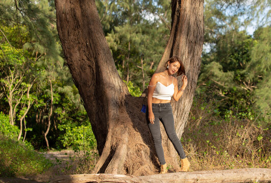 woman wearing sexy white tank top Wearing black tights and boots, .standing leaning against a large pine tree. .Tree Tunnel beside the beach. Large pine trees line up to form a tunnel background.