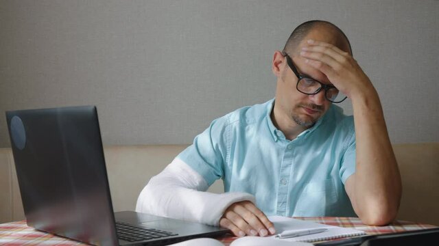 Man in eyeglasses with arm in plaster cast sitting in front of laptop and rubbing his bald head. Tired businessman working remotely from home after accident. Concept of stress