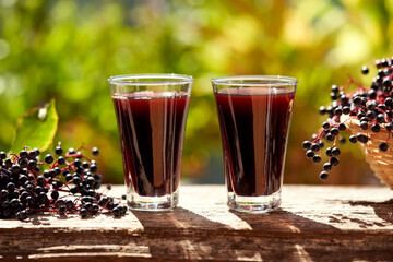 Two glasses of black elder syrup with fresh elderberries, outdoors with green bokeh