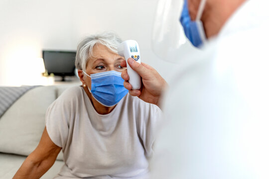 Cropped shot of a senior woman wearing protective face mask for safety against COVID-19 and getting temperature checked by male nurse at home during the day. Pandemic, COVID-19 virus outbreak concept.
