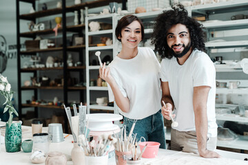Asian woman and afro american man with brushes in potter's wokrshop smiling.