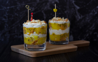 Two mango and passion fruit trifles with cream cheese and home made biscuit on a wooden cutting board. Dark background, side view