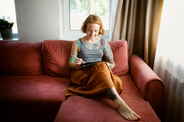 Obraz premium Cute red-haired girl artist with freckles draws on a tablet in a cozy living room real interior, sitting on a sofa in linen skirt , lifestyle