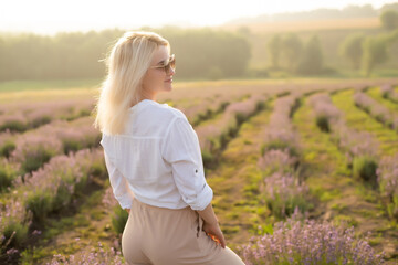 Fototapeta na wymiar Beautiful young healthy woman with a white dress running joyfully through a lavender field holding a straw hat under the rays of the setting sun