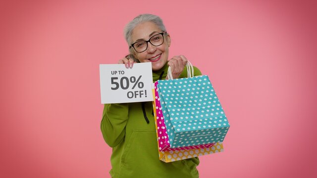 Senior Granny Woman Showing Shopping Bags And Up To 50 Percent Off Inscriptions Banner Text. Black Friday. Good Holiday Discounts, Low Prices For Shopping. Elderly Old Grandmother On Pink Background