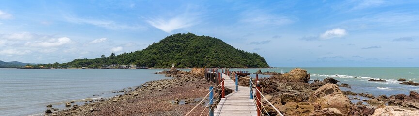 Fototapeta premium wide panorama view of seascape wooden bridge with rope rail to white pagoda on rock in blue sea, big mountain, and blue sky with cloud, Chedi Ban Hua Laem, travel destination in Chanthaburi, Thailand.