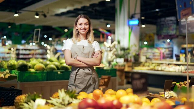 Portrait young woman worker in a Vegetable section supermarket standing smiling with arms crossed. Friendly pleasant female looking at camera in a fruit shop market. Employee in a work apron 