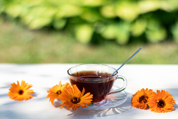 A transparent cup with a drink made from calendula flowers and calendula flowers on a light wooden surface on a natural background. Herbal tea with calendula, medicinal herbs.