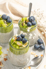 Healthy breakfast chia pudding with kiwi, blueberries and coconut slices, three portions in glass jars on a white table.