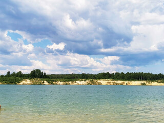 A clear blue lake with sandy shores surrounded by forest. Low rain clouds