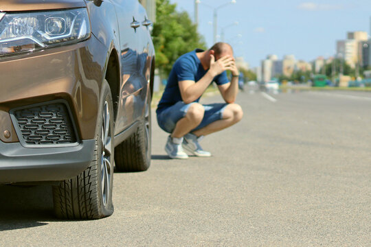 A Sad And Depressed Man Is Sitting Next To A Brown Car With A Punctured Tire.