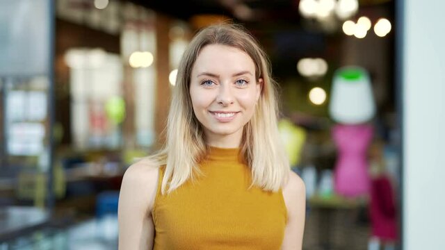 Close-up portrait of a young caucasian blonde woman standing and looking at the camera in a mall in a store or cafe on a blurred background. Happy female girl smiling in shopping center or supermarket