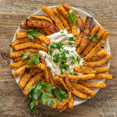 Baked pumpkin fries with tahini sauce and fresh parsley on white plate over wooden background.