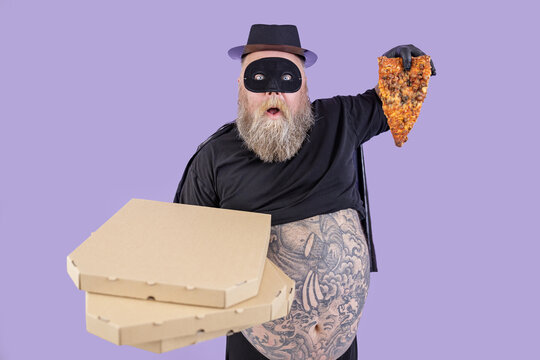 Shocked mature plump man in hero costume with large bare belly holds boxes and slice of pizza standing on purple background in studio